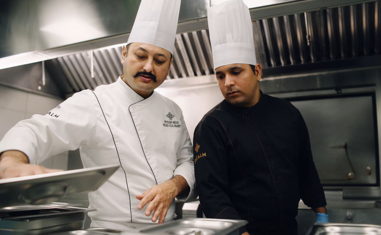 Two chefs in a commercial kitchen reviewing food preparation at a stainless-steel counter